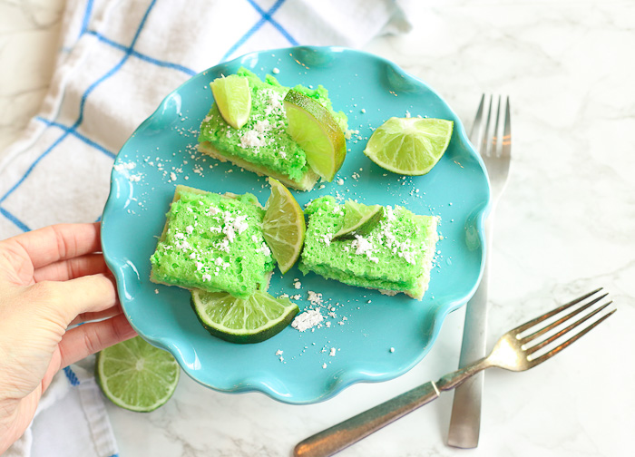 hand reaching for green gluten-free lime bars on a blue platter with powdered sugar and lime slices