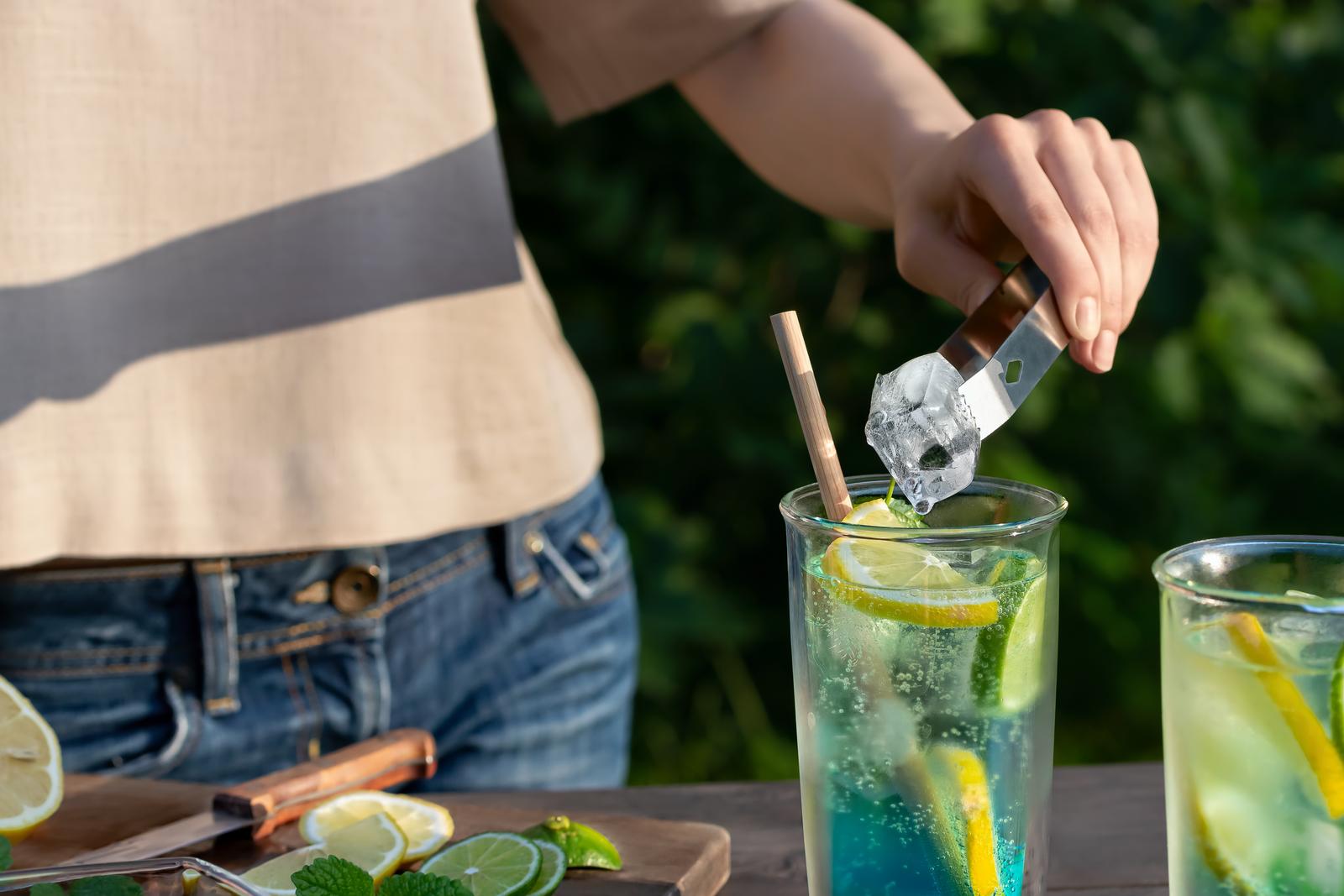 Girl makes summer refreshing cocktails blue lagoon. Close-up of adding ice cubes to glasses. 